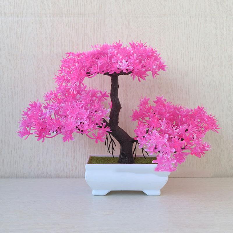 Artificial pink blossom bonsai tree with a curved trunk and dense floral canopy, placed in a glossy white ceramic pot against a neutral wood-textured background.
