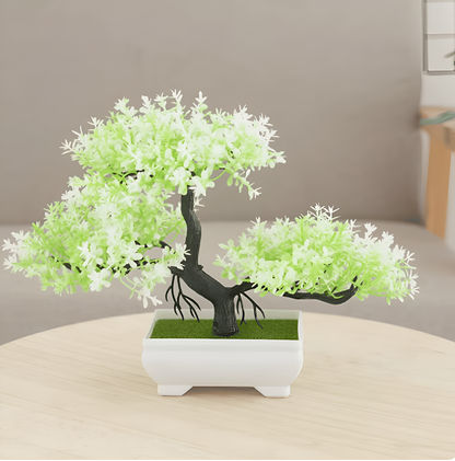 Artificial green blossom bonsai tree with realistic foliage and a dark curved trunk, displayed in a white ceramic pot on a light wood table in a cozy living room setting—perfect for indoor décor, desk styling, or zen-inspired home accents.
